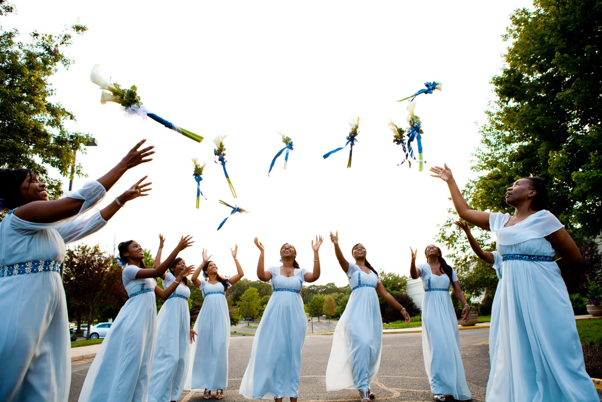 Bridesmaids in blue reach for bouquets thrown into a bright sky