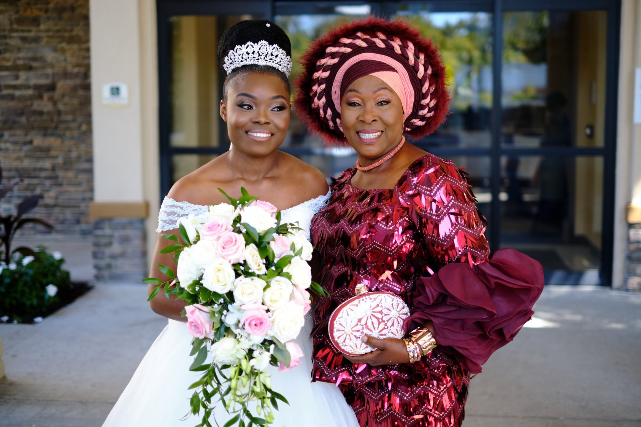 A bride with her bouquet beside her mother in a red gele