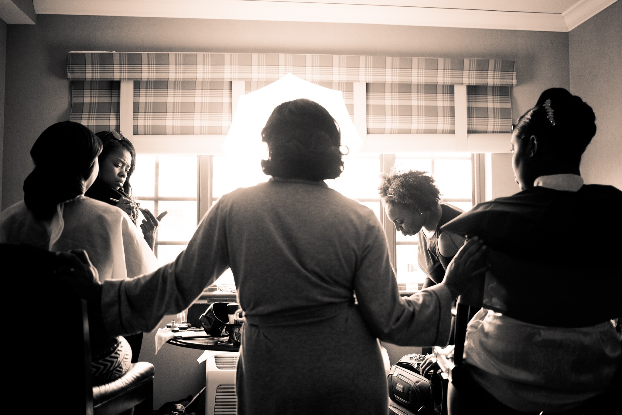 Women helping a bride dress by a bright window, in black and white